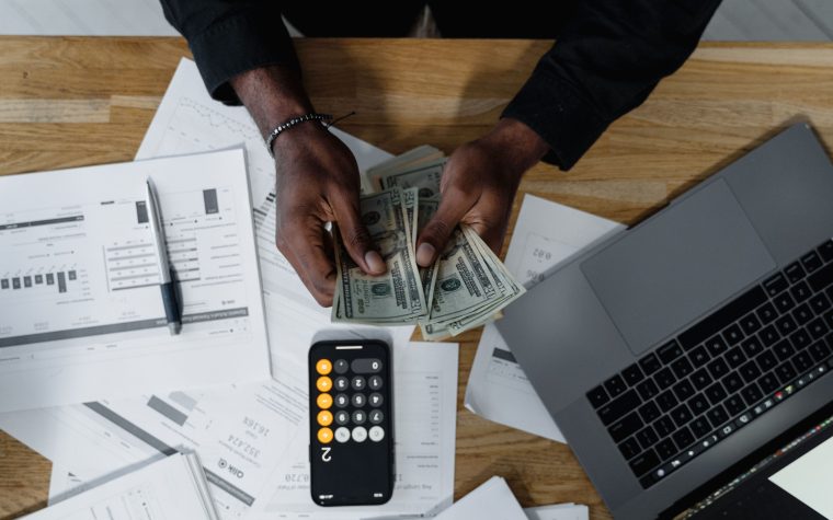 Person counting US dollars, using a calculator and laptop, with financial documents on a wooden desk. Une personne compte des dollars américains, en utilisant une calculatrice et un ordinateur portable, avec des documents financiers sur un bureau en bois.
