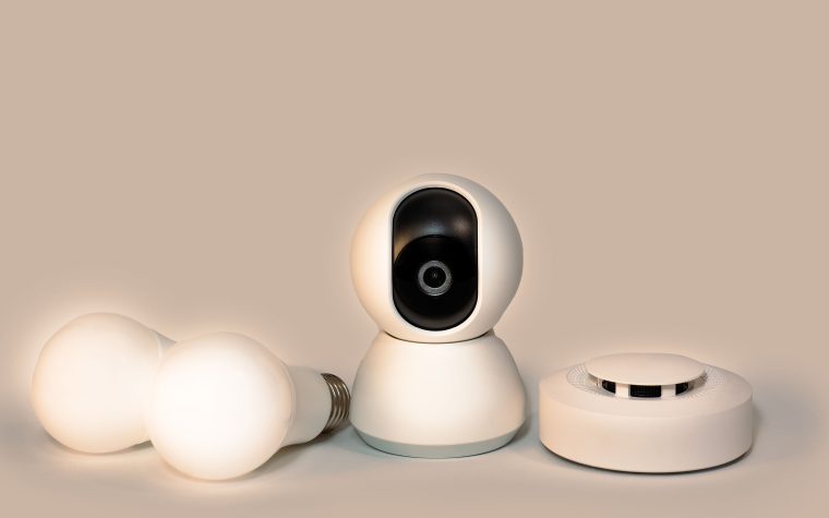 A collection of modern smart home gadgets including a CCTV camera, LED bulbs, and a smoke detector. Une collection de gadgets modernes pour maison intelligente incluant une caméra CCTV, des ampoules LED et un détecteur de fumée.